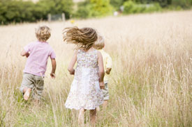 Three young children running through a grassy field toward the trees on a sunny day