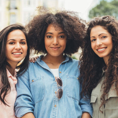 Three young women smiling outdoors, standing close together in casual clothing.