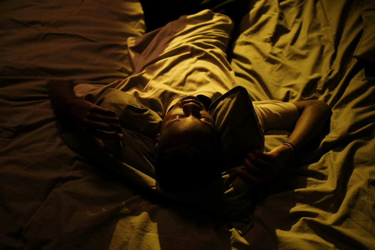 A person lies on their back in bed, illuminated by a warm, golden light, with their eyes closed and hands behind their head.