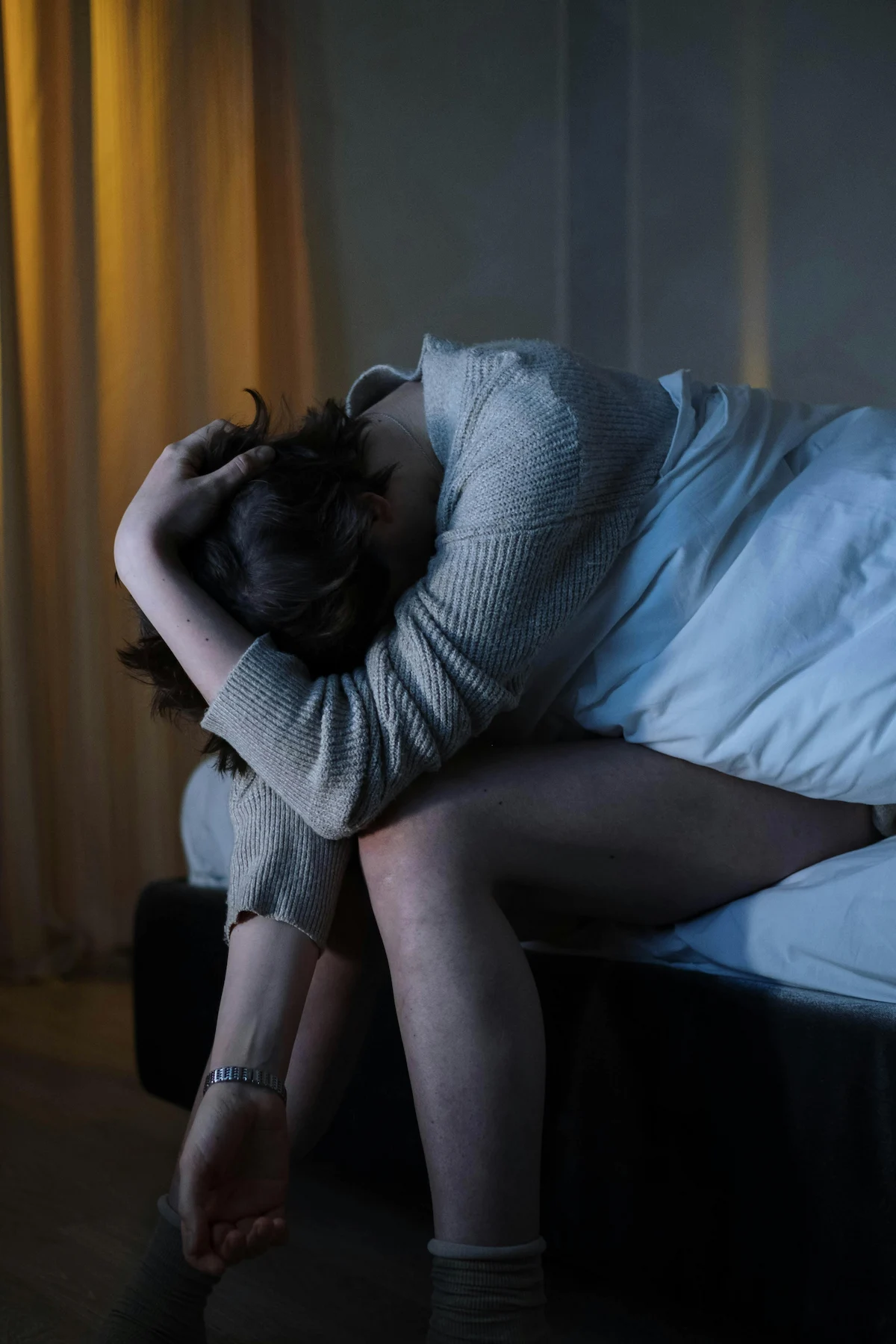 A person with head in hands tired sits on the edge of a bed in a dim room.